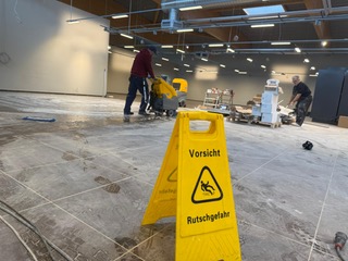 Ein Mitarbeiter der Herdegen Gebäudeservice GmbH reinigt mit einem Scheuersaugmaschine eine Baustelle. Ein Warnschild weist auf Rutschgefahr während der Nassreinigung hin.