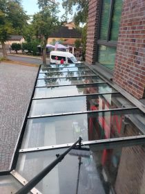 Ein Wintergartendach aus Glas wird mit einer langen Teleskopbürste gereinigt. Die Aufnahme zeigt die Anwendung moderner Reinigungstechnik auf einem Flachdach mit klaren Sichtscheiben und metallischen Trägern, im städtischen Umfeld.
